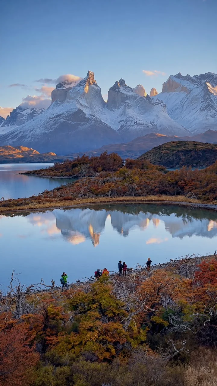 Un paisaje de montañas nevadas reflejadas en el agua, con un pequeño grupo junto a la orilla.